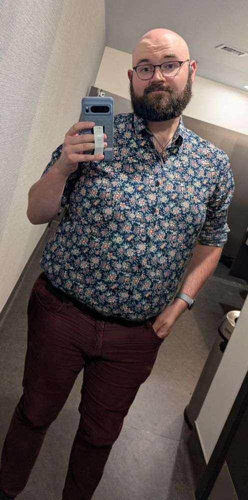 Bathroom mirror selfie of a bearded gay bear in a floral print button up and burgundy corduroys.