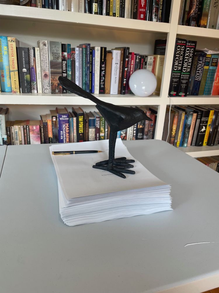 Thick manuscript on a desk in front of a bookcase. Atop the manuscript is a pen and a statuette of a crow with very large feet.