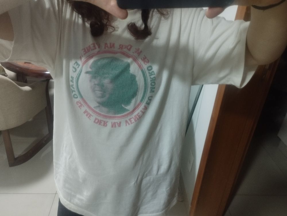 Uma pessoa vestida com uma camiseta branca com a foto de Jards macalé e escrito "se me der na veneta eu morro. Se me der na veneta eu mato"