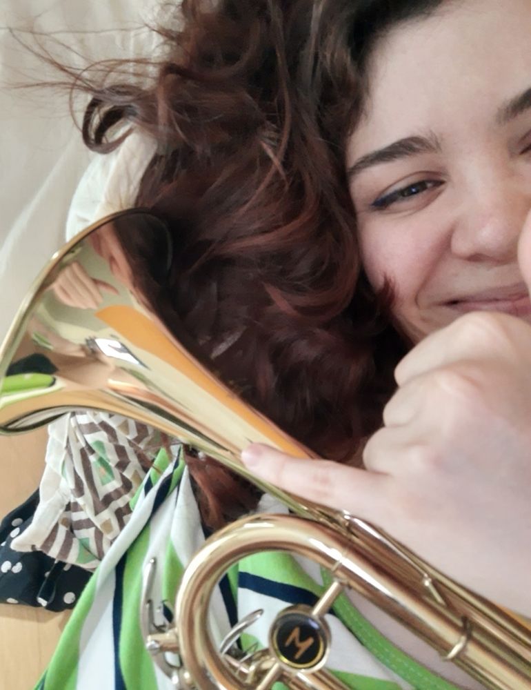 Uma mulher deitada sorrindo com seu cabelo solto e um trompete escorado em seu peito