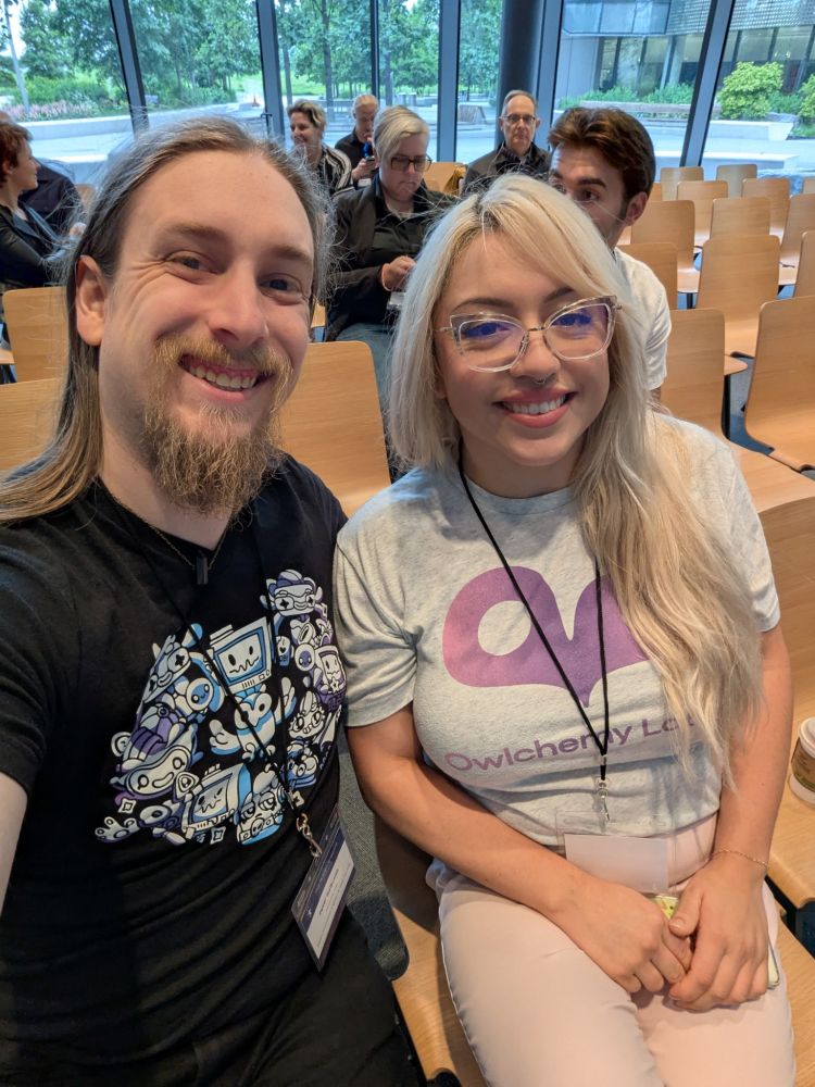 A selfie of Peter Galbraith and Jazmin Cano sitting in an auditorium. They are wearing Owlchemy Labs tshirts and smiling.