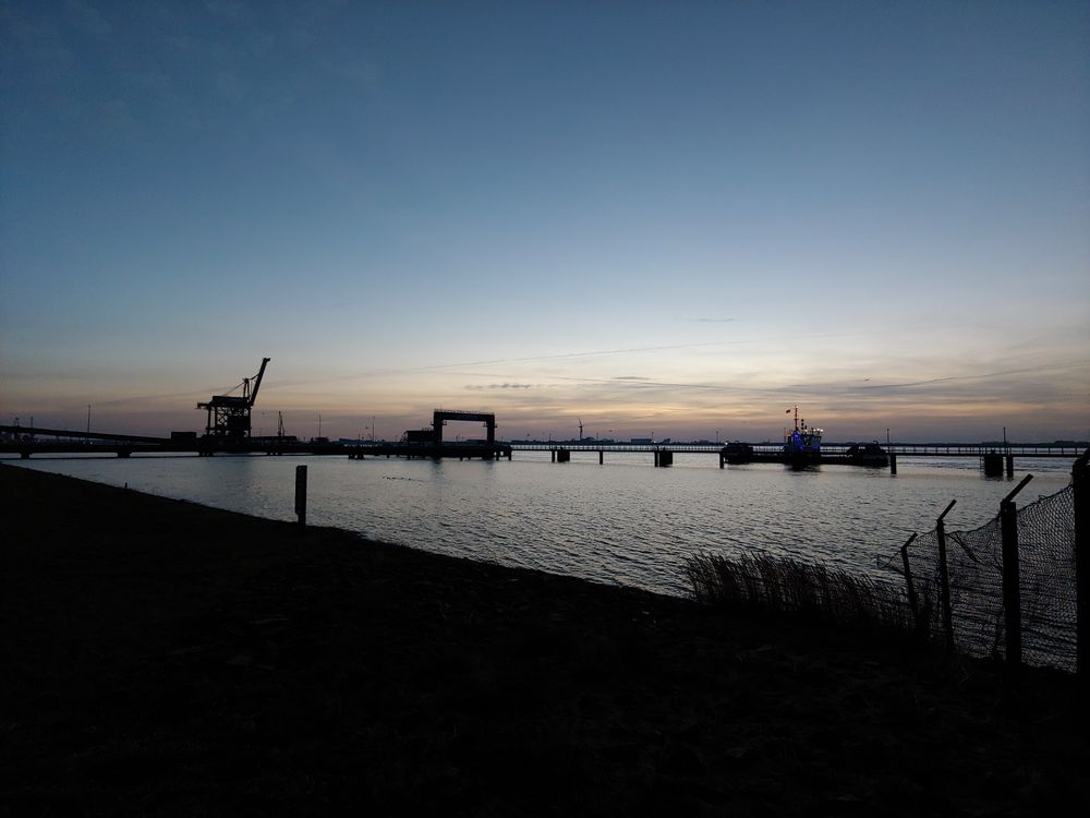 Dämmerungsaufnahme einer Hafensilhouette mit einem Kran, einer langen Pier und einem beleuchteten Schiff vor einem klaren, farbigen Himmel über leicht bewegtem Wasser.