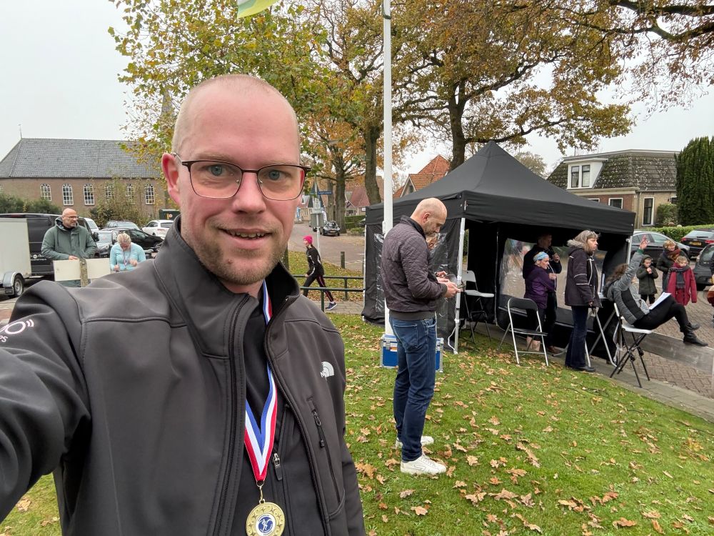 Picture of me, bald white guy wearing a black jacket and a medal, standing on grass, in front of a small tent where the finish line is for a running event.