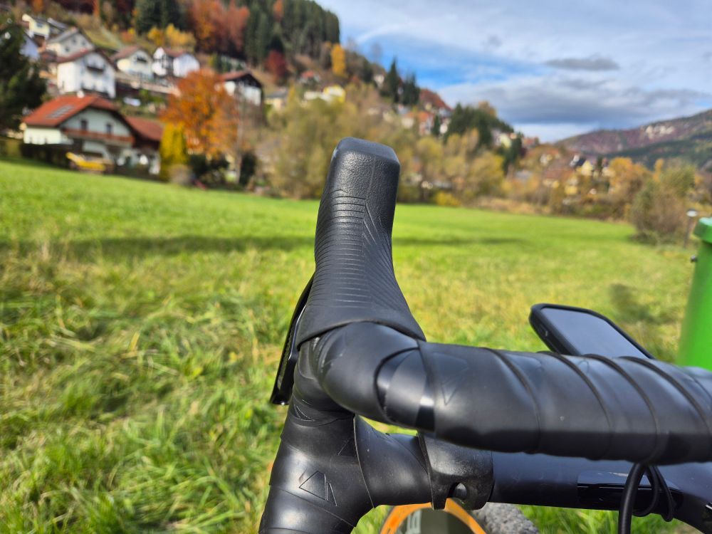 Halber Gravelbikelenker im Fokos, dahinter Tallandschaft mit Wiese, Häusern und Wolken am Himmel.