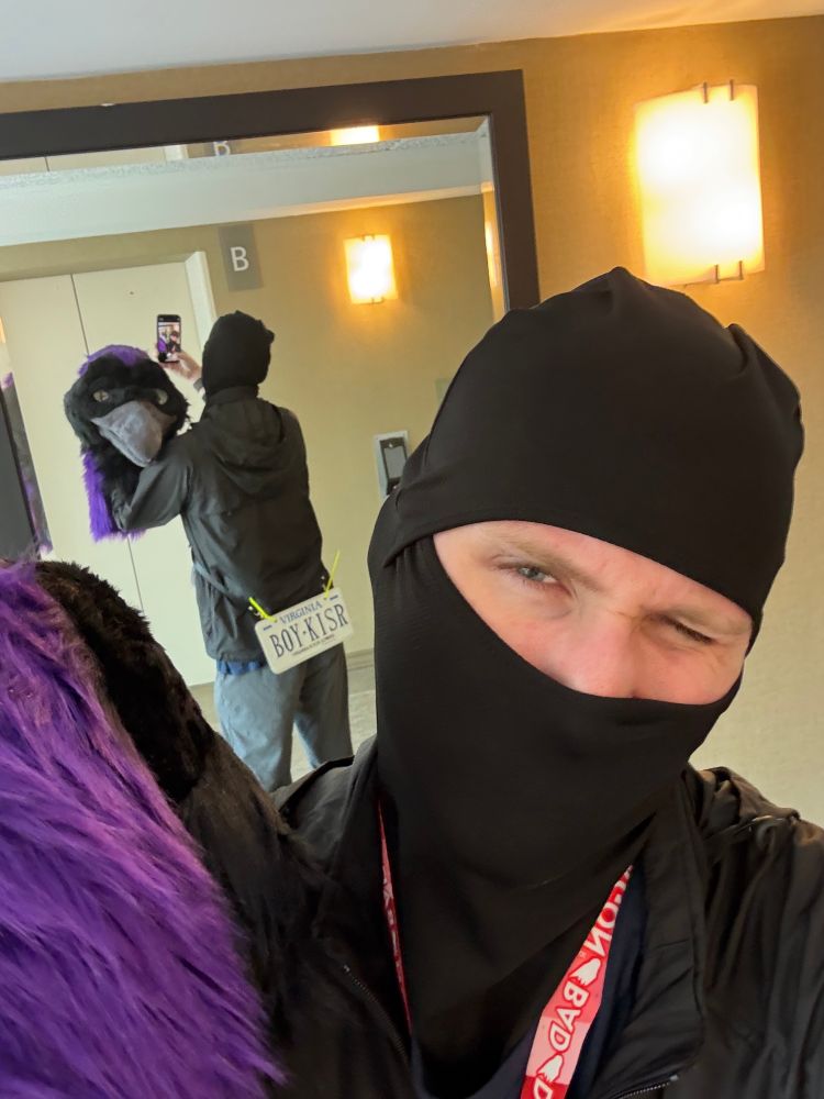 Mirror selfie of fursuiter. Mirror in background shows their fursuit head, a crow with purple accents, and a license plate strapped to their ass that reads “BOY-KISR”