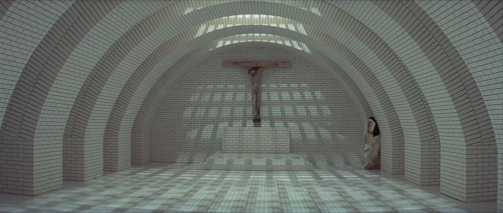 Still from The Devils (1971): A pale tiled rounded room with a crucifix at one end, and a nun tucked into a corner.