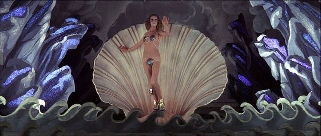 Still from The Devils (1971): a stage with cutouts of waves, and a performer imitating The Birth of Venus with a clamshell as a backdrop.