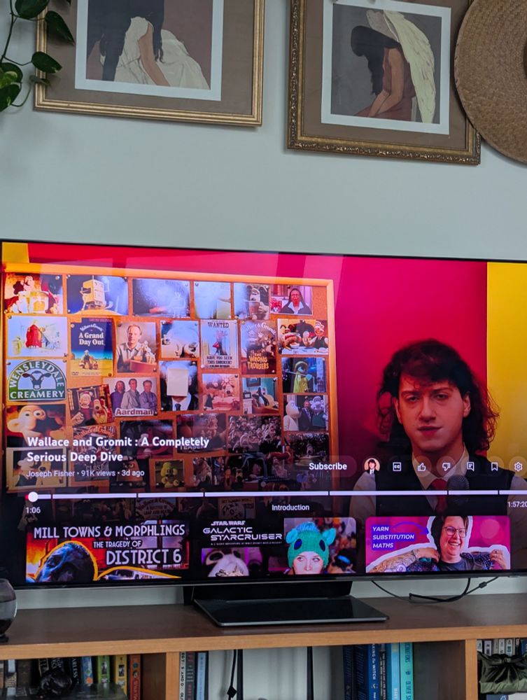 A TV screen paused on a YouTube video titled "Wallace and Gromit: A Completely Serious Deep Dive", by Joseph Fisher. The video is 2 hours long. The paused screen shows a young white man (Joseph Fisher) dressed as Wallace in a white shirt, green knit vest, and red tie holding a mug that is shaped like Gromit's head. The rest of the screen is filled with a corkboard covered in  Wallace and Gromit-related pictures. 