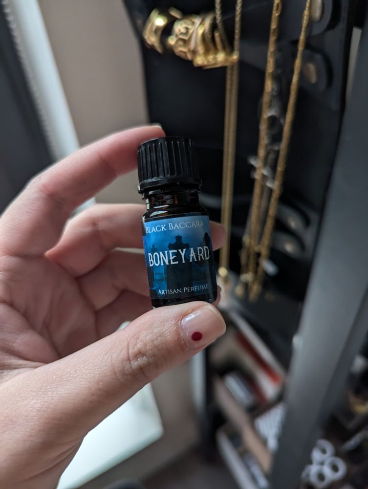 A hand holds a small 5ml apothecary bottle of perfume oil. The label on the bottle says "Boneyard" in large letters, "Black Baccara Artisan Perfume" in smaller letters, on a background picture of black gravestones against dark blue fog. 