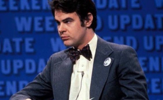 Photo of Dan Aykroyd in front of a blue Weekend Update backdrop. He is wearing a dark gray suit jacket with a WU pin on the lapel, a white shirt, and a bow tie. He looks very serious and very handsome.