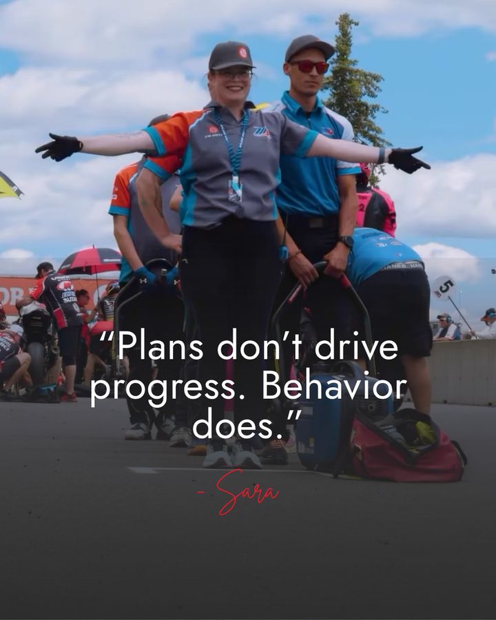 Sara stands on the motorcycle race grid, with her arms out (waiting to catch the bike). She stands next to a teammate holding a bike stand. She's in a blue, orange, and grey team uniform and baseball cap. She's smiling, broadly.