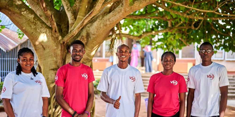 Five volunteers in Nigeria are standing outside in front of a tree. They are wearing white and pink VSO t-shirts, smiling to the camera. They are part of the National Youth Engagement Network in Nigeria.
