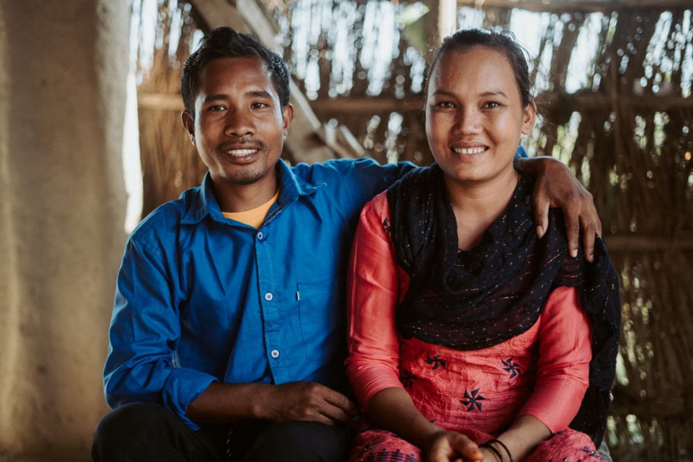 Two people in Nepal smiling and sitting close together in a rustic setting with a thatched background.
