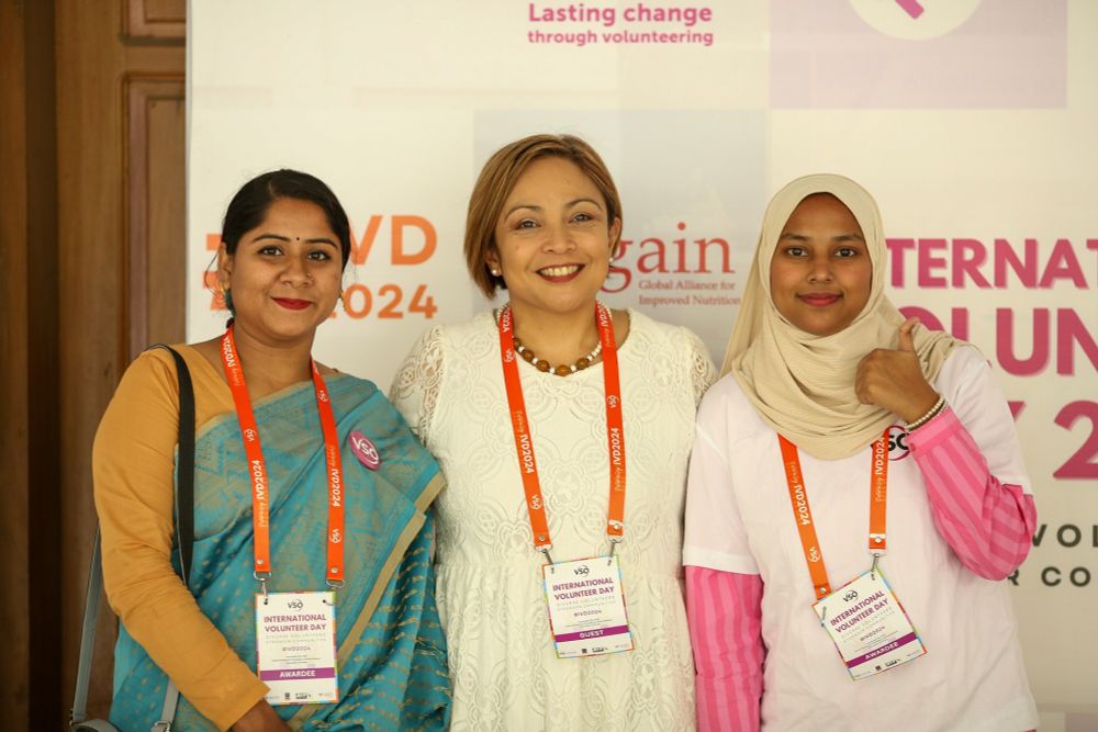 Three women are stood in front of a branded selfie wall wearing orange lanyards with name tags on them.
