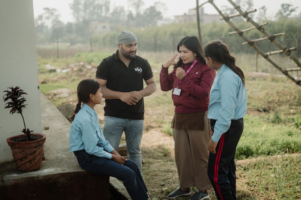 In Nepal, two VSO volunteers are stood with two schoolchildren. They are in an outdoor setting with lots of greenery behind them. They are all engaged in a conversation.