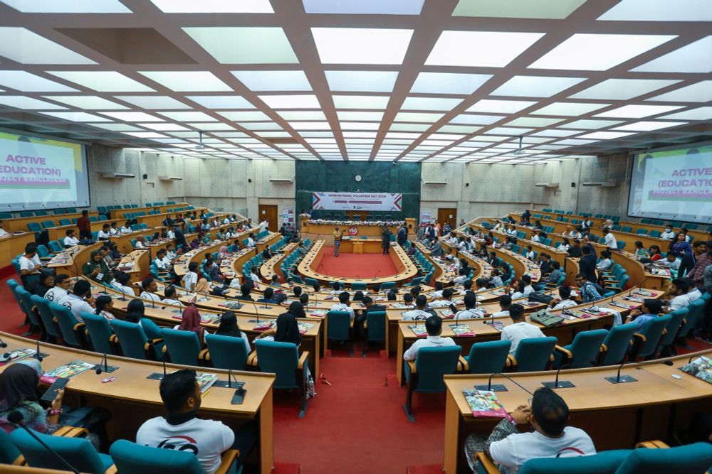 A conference hall with tiered seating and desks wrap around the room. There are volunteers sat in seats looking forward to a volunteer speaking. There are posters on the walls with VSO logos on them.