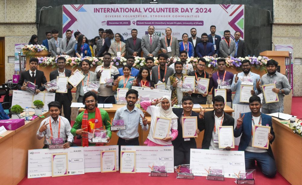 Three rows of VSO volunteers are smiling and holding certificates that are acknowledging their achievements from the past year. They are all smiling in front of a VSO branded sign that says 'International Volunteer Day 2024'