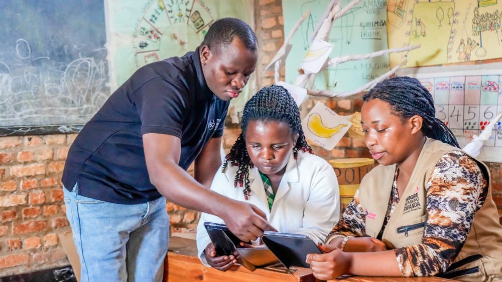 VSO volunteer and award winner Celestin is stood wearing a black shirt and denim jeans. He looks to be in a classroom setting, and is leaning down pointing at tablets held by two women. One is wearing a white jacket and the other is wearing a beige gilet-styled overcoat.