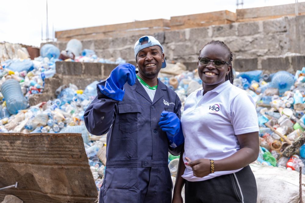 Two workers smiling at a recycling site, one wearing a blue jumpsuit and PPE equipment, the other in a white polo shirt with the 'VSO' logo. Both are standing in front of a pile of compressed plastic waste.