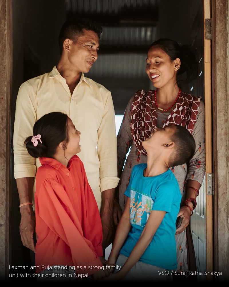A family of four are stood in the doorframe of their home, all looking at one another. Laxman (top left) and Puja (top right), stand behind their two young children.

There has been text added on top of this image which reads: Laxman and Puja standing as a strong family unit with their children in Nepal.

Photograph by; Suraj Ratna Shakya. 