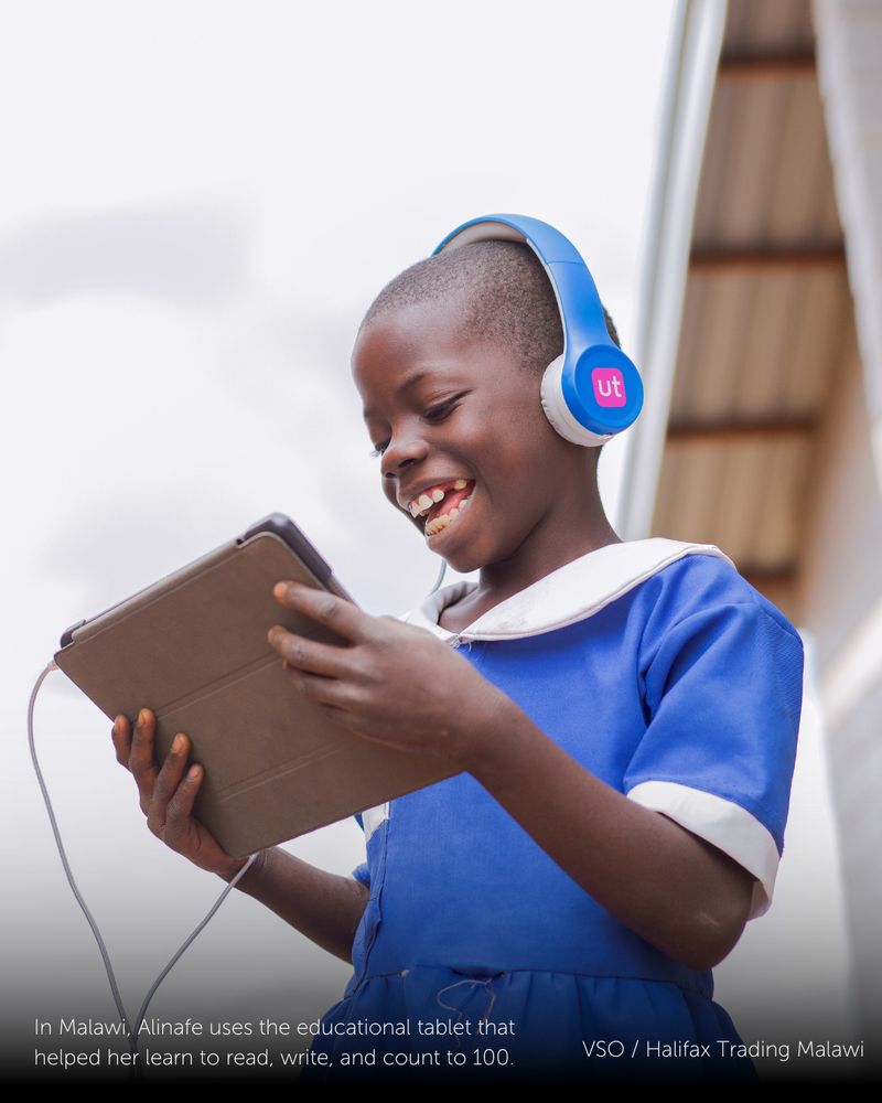 A young girl in Malawi, wearing a bright blue uniform with a white collar, is wearing headphones while holding a tablet. She is smiling as she looks down to what is on the screen.

Text at the bottom of the image reads: "In Malawi, Alinafe uses the educational tablet that helped her learn to read, write, and count to 100. - VSO / Halifax Trading Malawi."