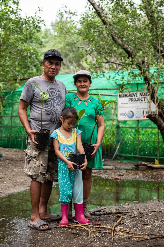 In the Philippines, a father and mother stand with their small daughter. They are located in an outdoor environment holding trees that are ready to be planted.