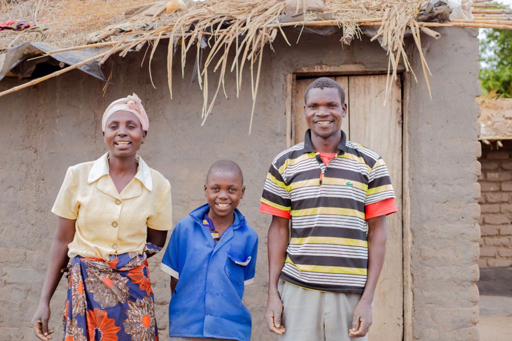In Malawi, Thoko is stood between his mother and father outside of their home.