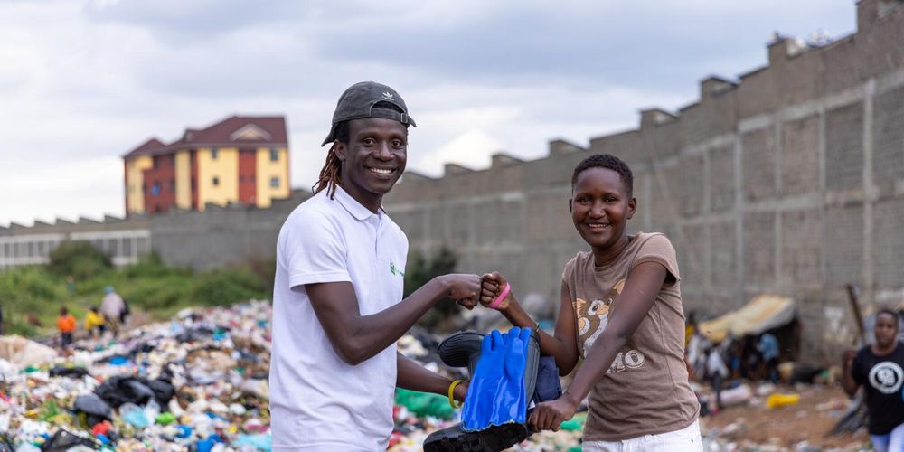 VSO volunteer Lawrence is wearing a white t-shirt and cap. He is stood in a waste dump site in Kenya with a woman who he is supporting through VSO's From Waste to Work project. She is wearing white trousers and a brown t-shirt.
