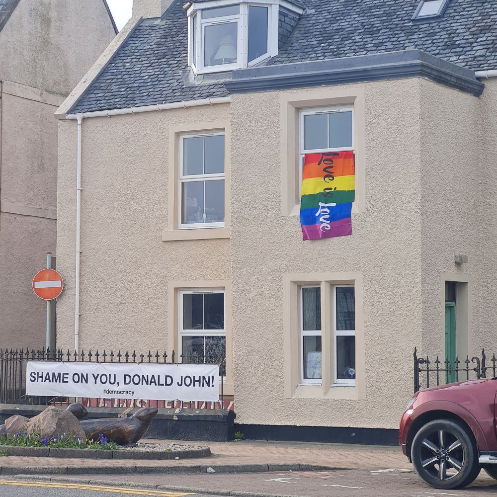 White washed two story  house on the harbour- you can't quite make out it's a gift shop/gallery. On the railings outside, black print on a white banner- "SHAME ON YOU DONALD JOHN #democracy" and upstairs, right hand window, a rainbow flag with love is live on it. 