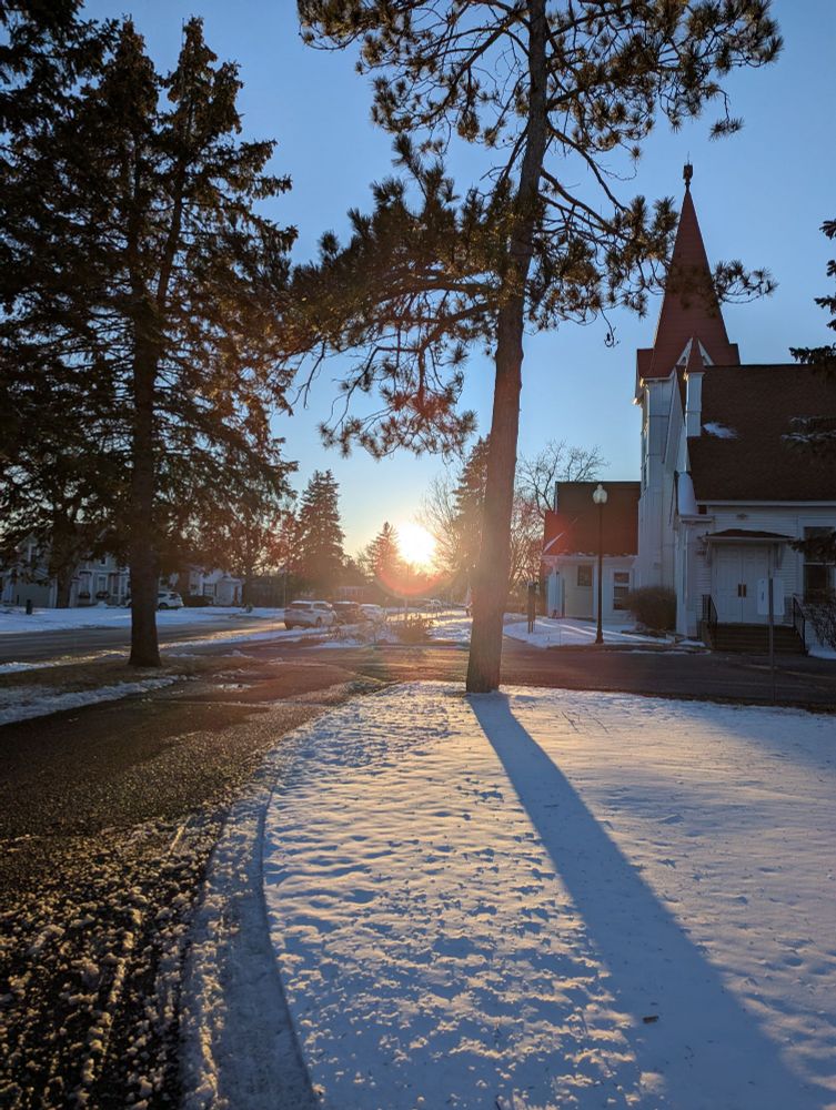 The sun setting between trees next to a church.