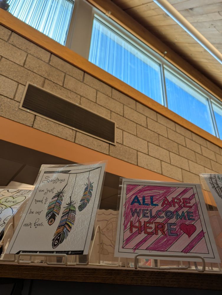Art on a library bookshelf stating, "Sometimes we just need to be our own hero," and "All are welcome here."