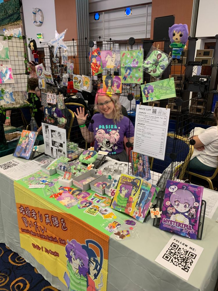 Pink haired lady sitting behind a convention table containing comics, stickers, etc.