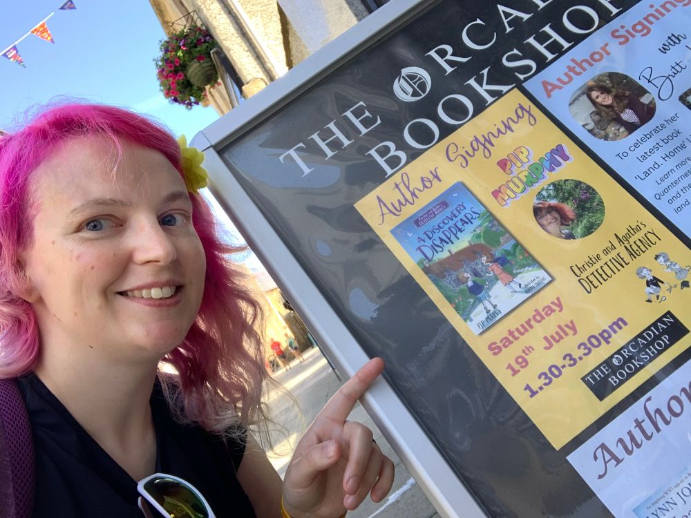 A pink haired lady pointing at a yellow poster for an Author Signing with Pip Murphy