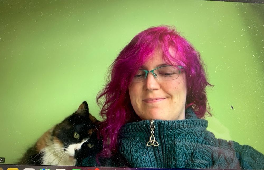 A calico cat nuzzling against an amused-looking pink haired lady wearing turquoise glasses, a greeny-blue zip-up arran sweater with a Celtic looking metal zip. There is a light green wall behind. It is a photo from a Zoom meeting.