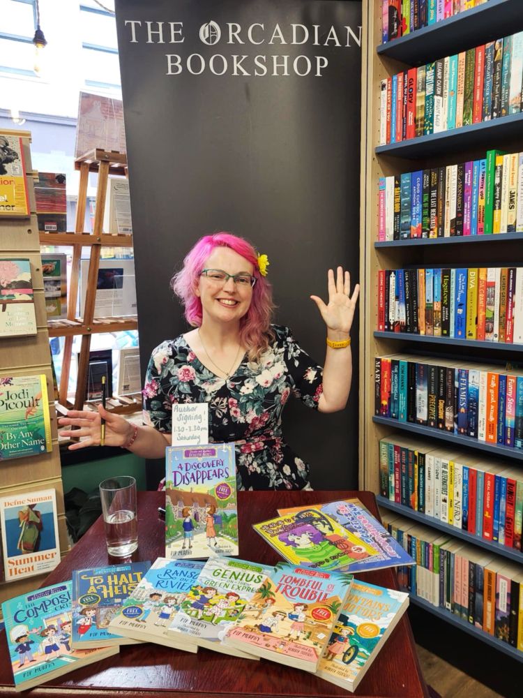 A pink haired lady with a yellow flower in her hair sitting behind a table full of Christie and Agatha Detective Agency books with a banner reading “The Orcadian Bookshop”.