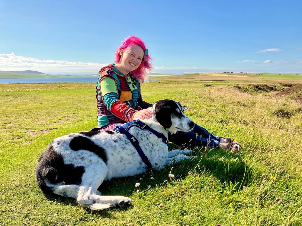 Black and white dog with pink haired lady in a multicoloured hoodie sitting on the grass on a fine day with a blue sky.