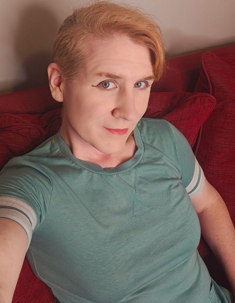 A redheaded transfeminine person, Sarah Miranda, is leaning back on a red couch 