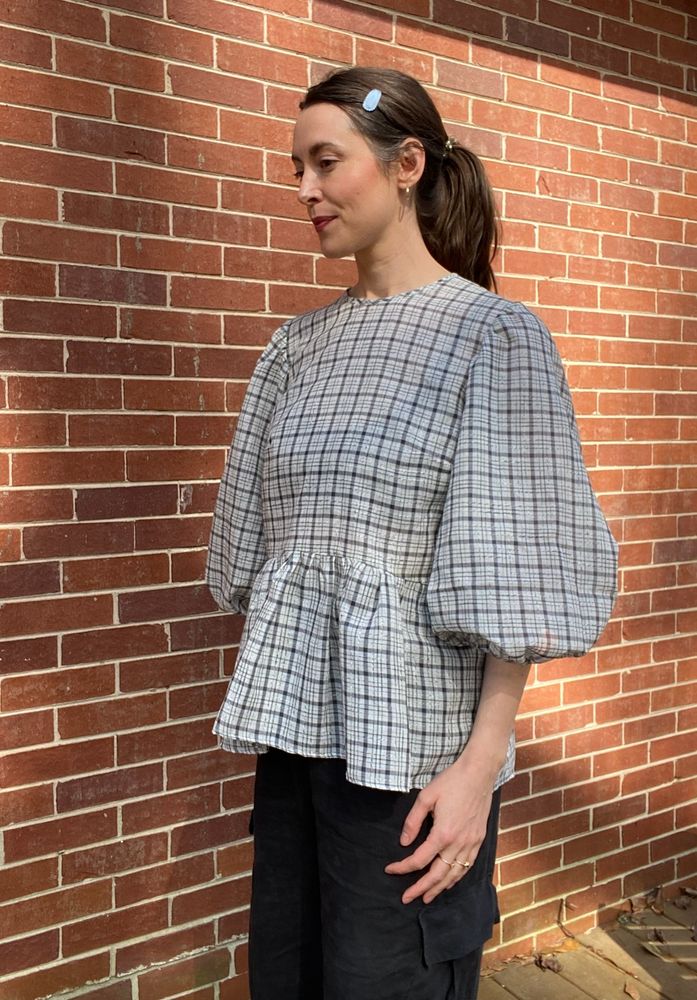Pictured is a white woman in her thirties wearing an black and white plaid puff sleeve 3/4 length peplum shirt in an organdy fabric and black linen cargo pants