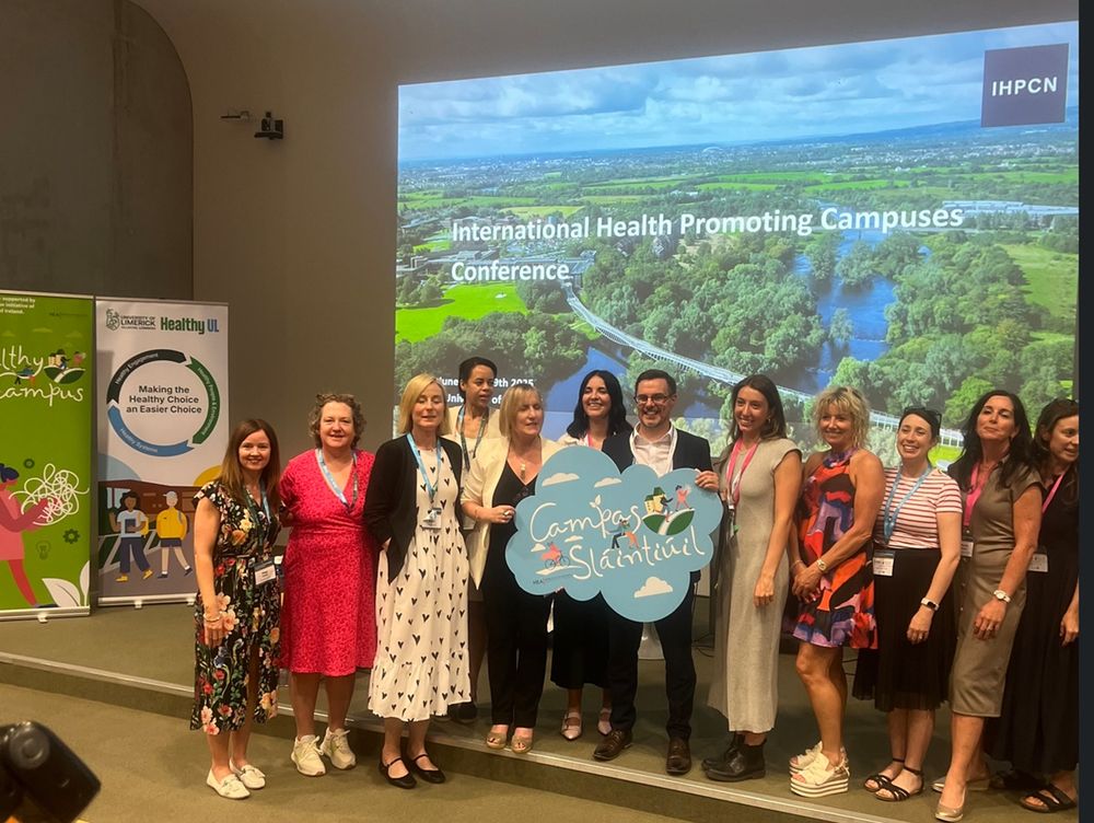 10 women and 1 man at IHPC Conference screen backdrop, one holds sign for Campas Slántiúl
