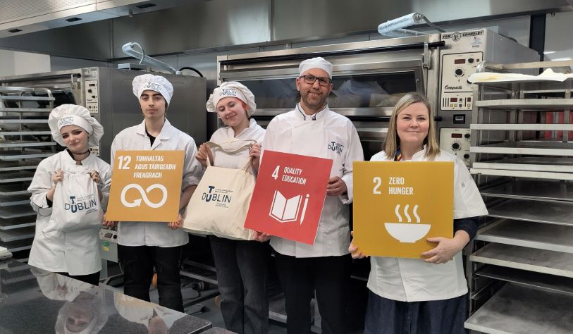 3 white females and 2 white males hold up SDG signs 2,4 and 12 & reusable bread bags in bakery uniforms in kitchen.