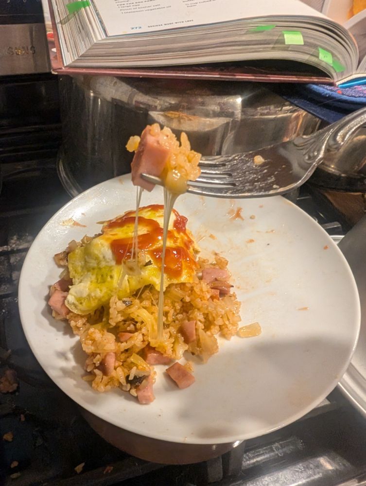 A fork holds a bite of fried rice with ham and a long cheese pull dangling down to the plate