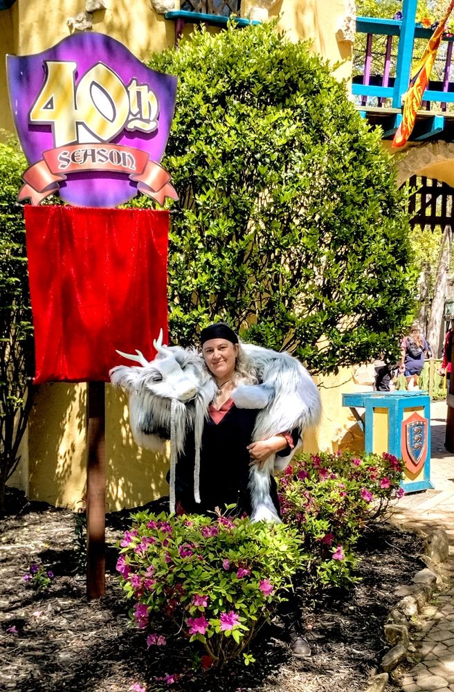 Kate stands with her dragon puppet of White and light blue at the front of the Georgia Renaissance Festival, standing near the banner for their 40th season