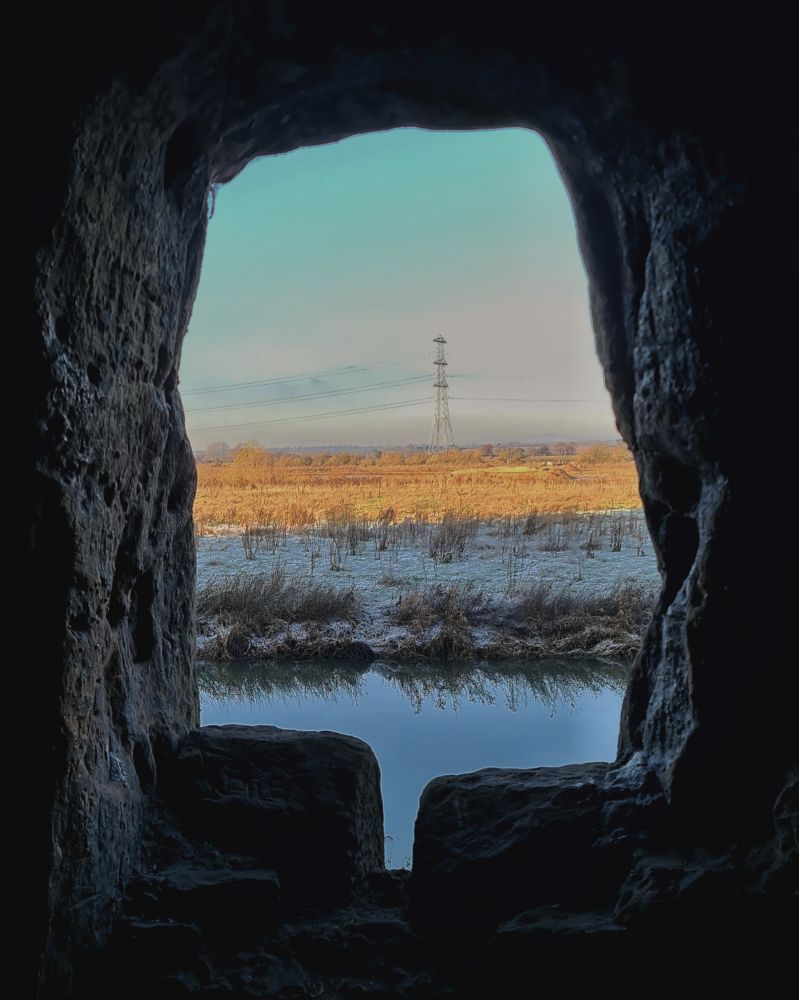 Portrait colour photo of an irregular hole of a window carved in solid rock in a cave. Through the window is a winter scene in orange, white and pale blues. Still water in front, then a flat frozen field, half white in the shade and half lit orange in late afternoon sunlight. At the back in the distance is an electricity pylon and cables against the sky which is white with a touch of pink lower down, and blue at the top. 