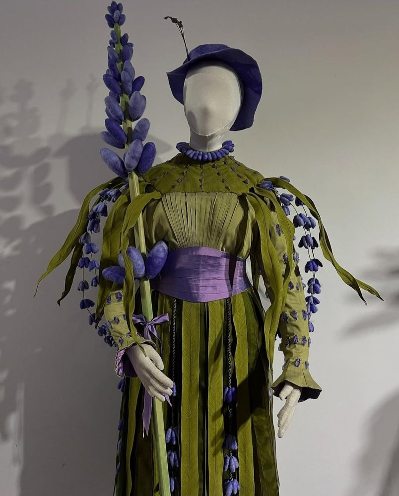 Portrait colour photo. A mannequin stands against a white wall. It holds a huge spear of lavender. The dress it wears is in a Victorian style, in olive green. It dobs a purple hat with a sprig of lavender on top, a lavender flower ruff. There are lavender sprigs bursting out of the shoulders and draping down the long green sleeves. The waist band is purple, and there are lots of other little lavender and purple accents and details. 