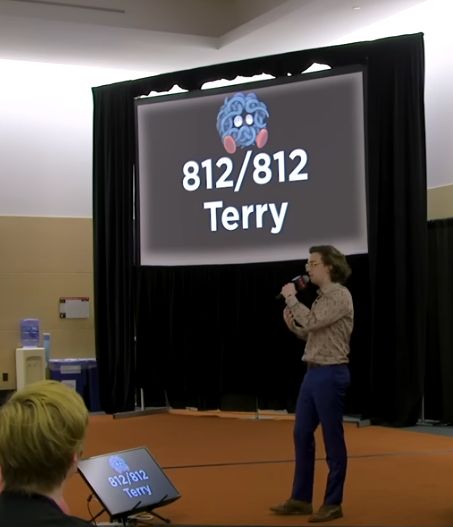 A screenshot of the Perfect Pokerap from Polygon, with YouTube creator and comedian Brian David Gilbert singing at a gaming convention with a picture of the Pokemon Tangela behind him.