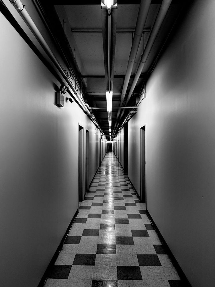 A black and white photograph of a long building corridor lit by fluorescent lights with a tile floor. The corridor is so long the end almost vanishes into the distance. Doors line both walls and the corridor is empty save for a tiny figure that can just be made out near the far end.