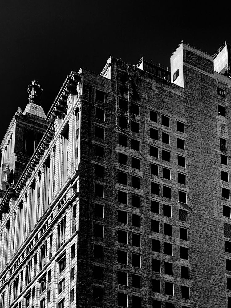 A black and white photograph of a high-rise building. The front of the buildings is soaked in sunlight and finished with elegant details framing the windows, while the shadowed side of the building is a utilitarian brick facade with stark rectangular windows.  