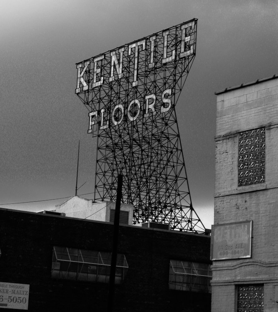 A black and white photograph of low-rise industrial buildings, one of which has a tall metal framework its roof, atop which are letters spelling out "Kentile Floors"