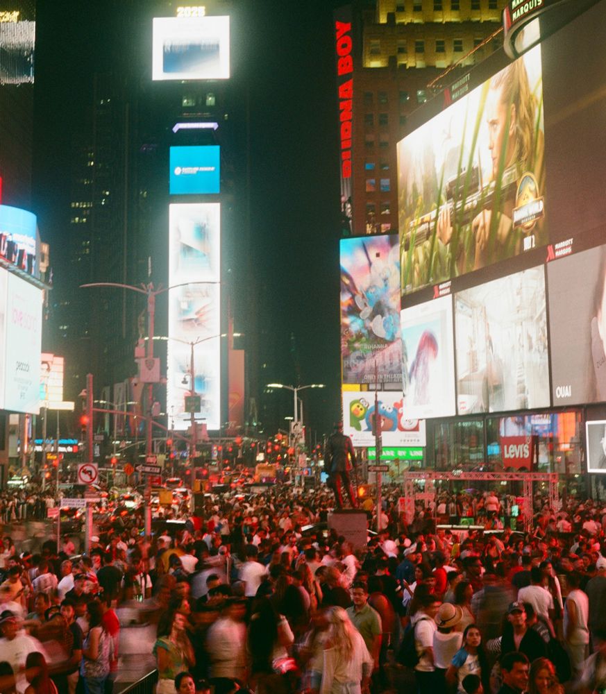 A photograph of a city intersection at night: towering bright screens on the surrounding buildings illuminate crowds filling the streets below. Some members of the crowd are motion blurred while others are sharply distince. 