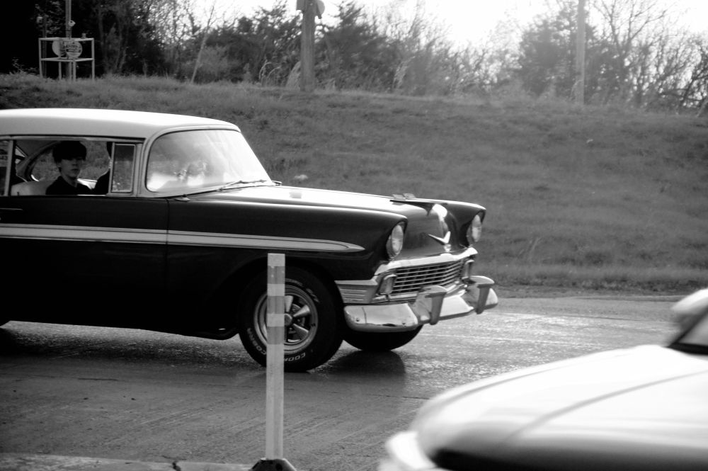 A black and white photograph of a vintage car stopped at a traffic intersection in front of a grass-covered bank of earth. A young man is looking toward the camera from the passenger seat through an open window: most of his face is shadowed, but his visible expression reflects suspicion.